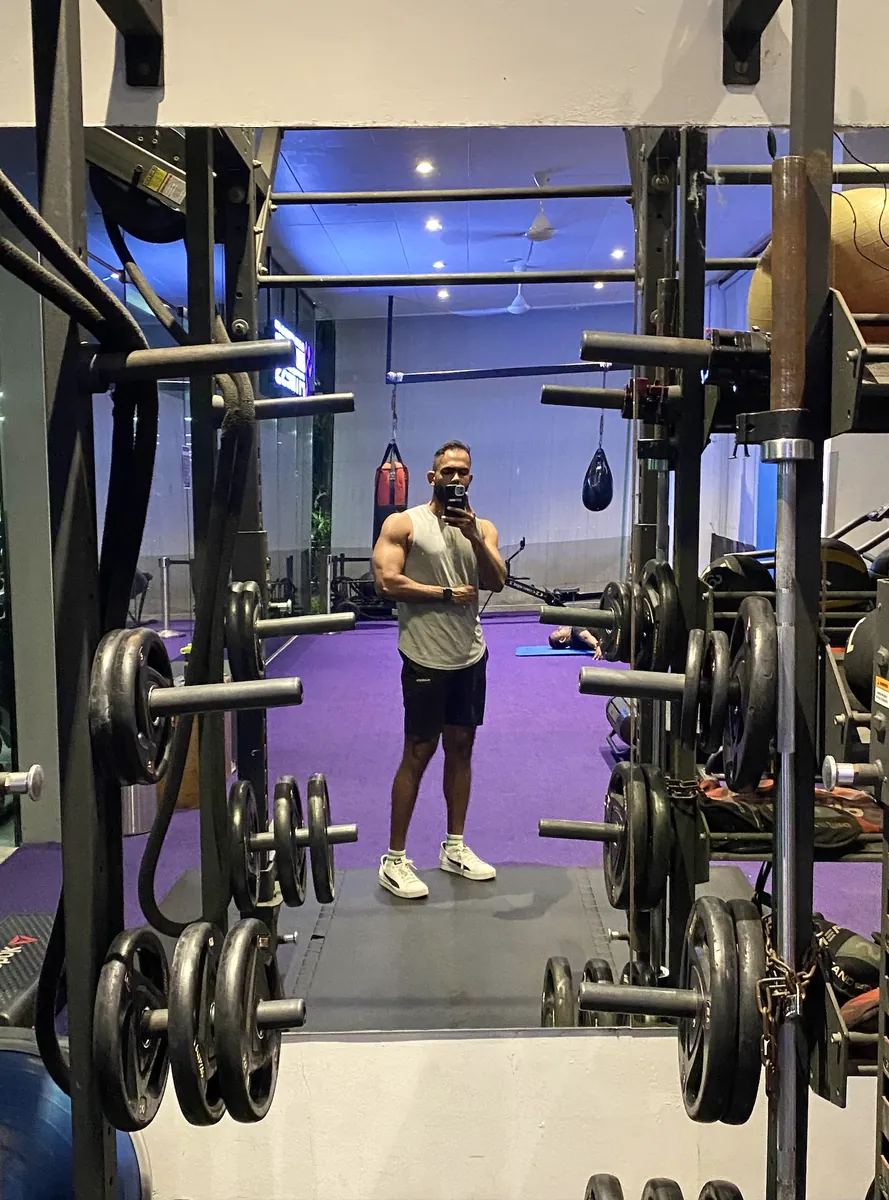 Anuja in a gym, taking a mirror photo between two loaded squat racks.