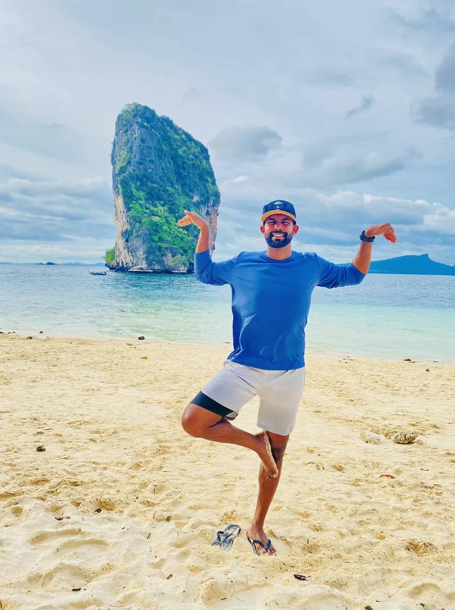Anuja balancing on one leg on a white-sand beach, a limestone karst rising out of turquoise water behind him.