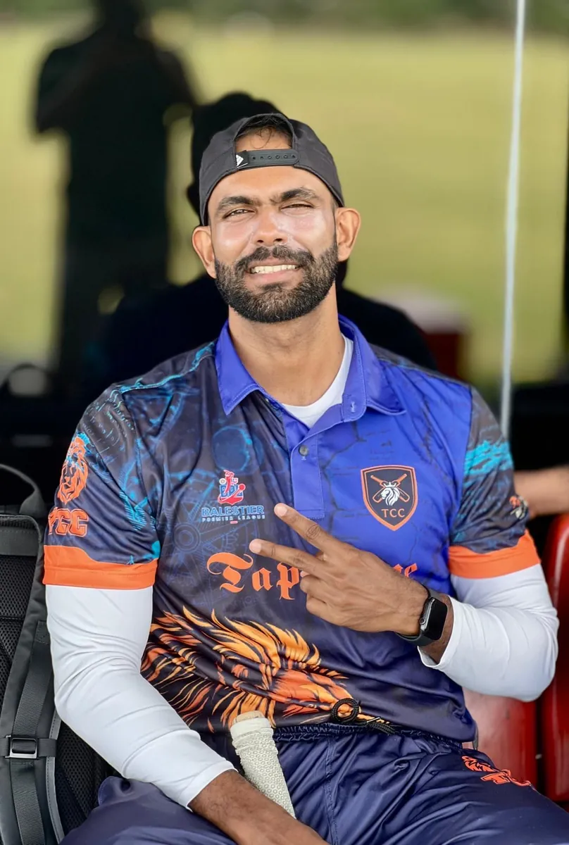 Anuja in a Tigers Cricket Club shirt and backwards cap, sitting by the boundary with a bat between his knees.
